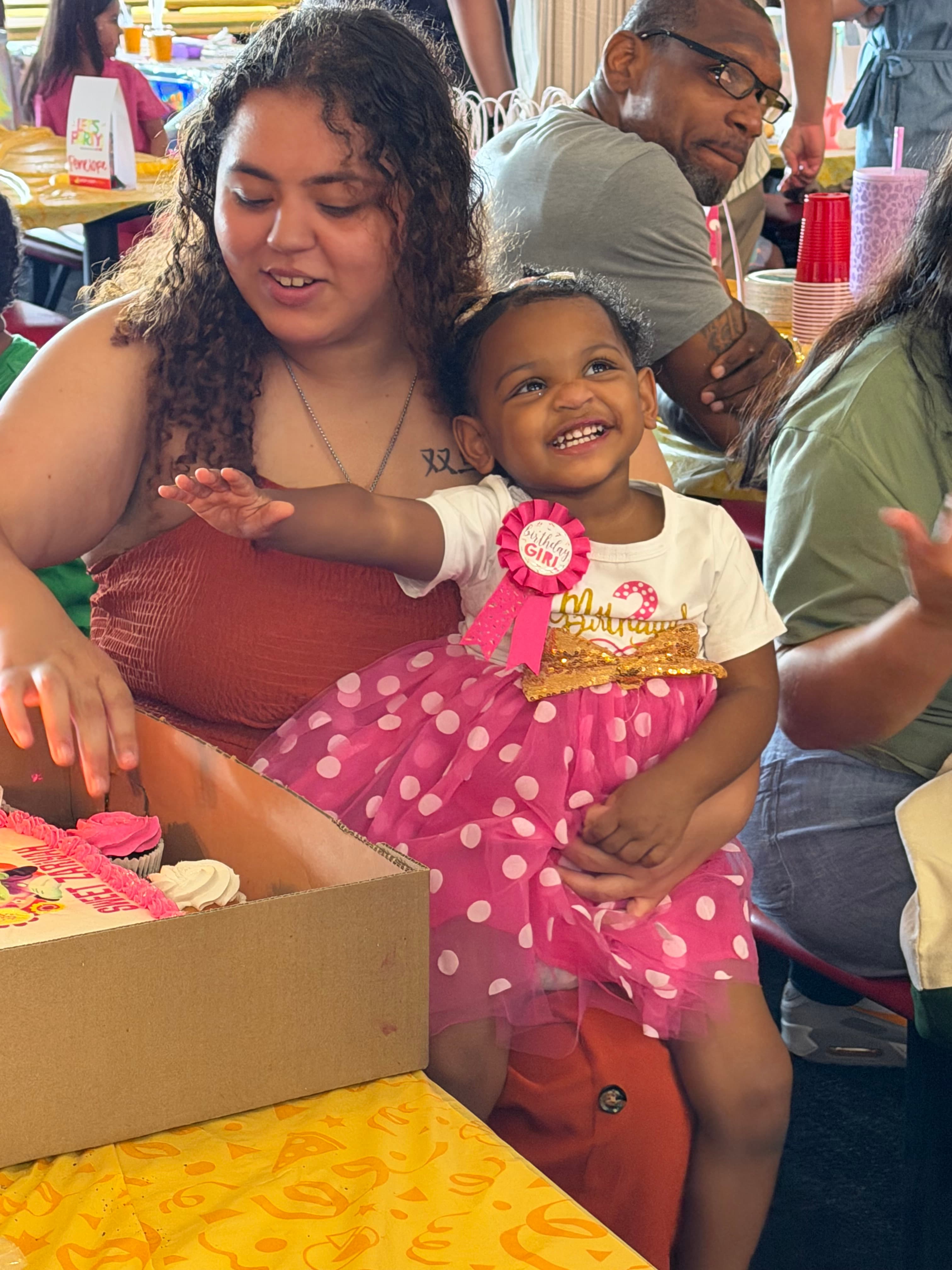Smiling toddler in pink polka-dot tutu sits on a woman's lap near birthday cake.